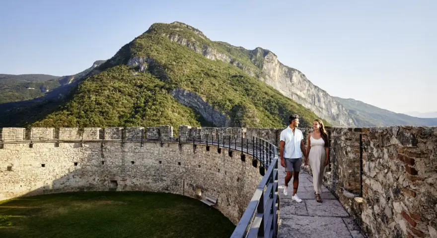 Coppia passeggia su un bastione di un castello con montagne verdi sullo sfondo. Un'esperienza da vivere nel Trentino da scoprire.