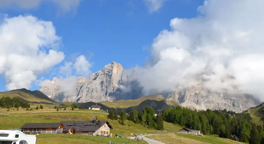 Panorami mozzafiato per escursioni a Passo Rolle. Rifugi alpini e vette imponenti sotto un cielo azzurro con nuvole bianche.