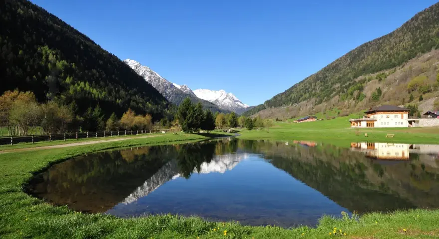 I laghetti di Vermiglio riflettono montagne innevate e un cielo azzurro. Un edificio moderno si specchia nell'acqua calma.