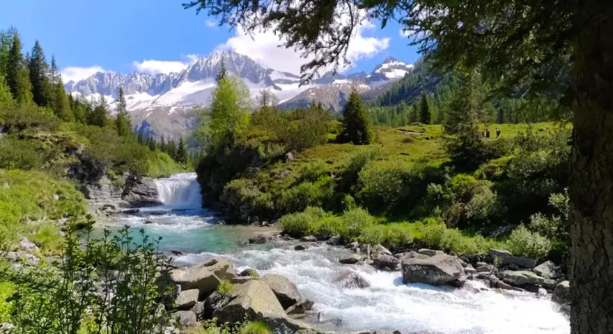 Un torrente impetuoso scorre tra rocce e vegetazione lussureggiante nel **Val di Fumo**, con montagne innevate sullo sfondo.
