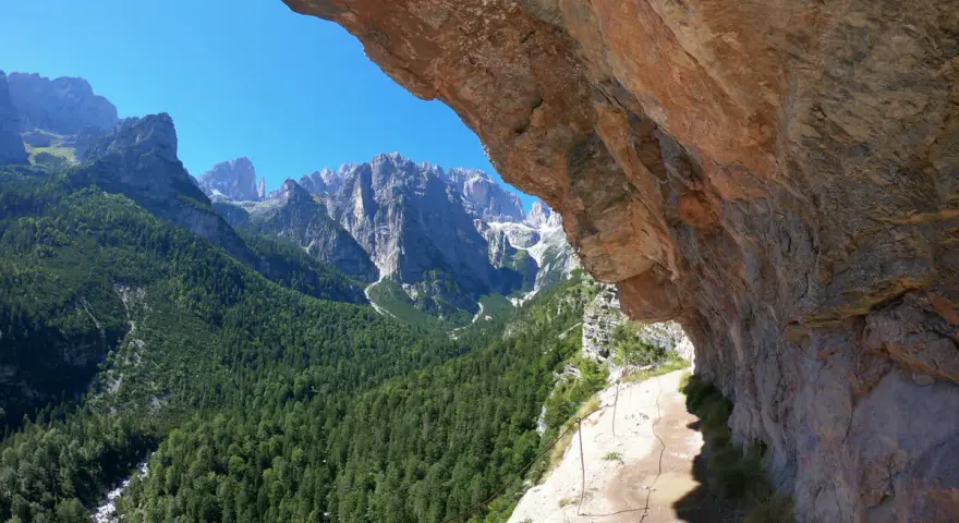 Sentieri panoramici vicino al Lago di Molveno, con un sentiero che costeggia una parete rocciosa e una vista mozzafiato sulle montagne e una cascata in lontananza.