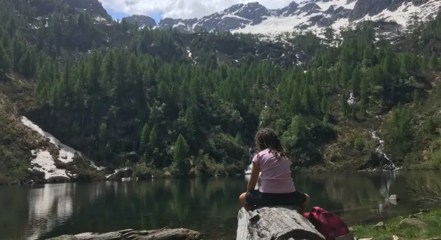 Bambina seduta su un tronco guarda il lago di Barco, circondato da montagne innevate e foreste di conifere.