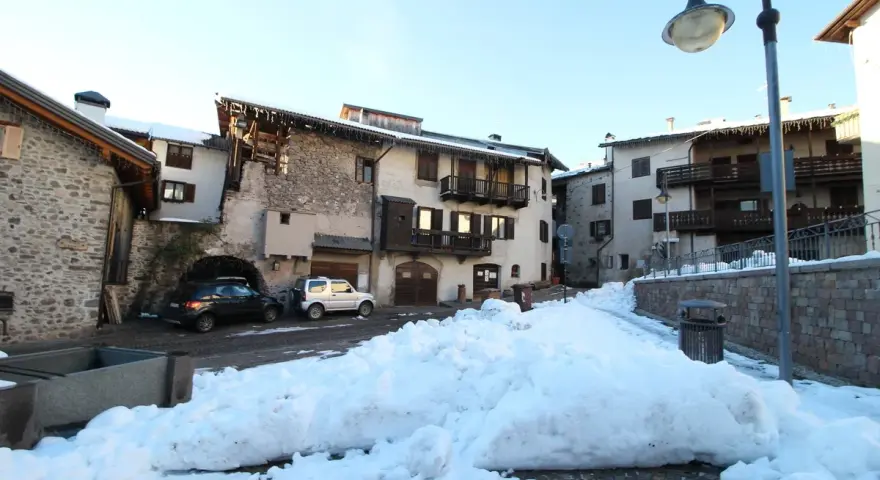 Paesaggio innevato a Miola di Pinè, con case in pietra e legno, auto parcheggiate e un cielo azzurro.
