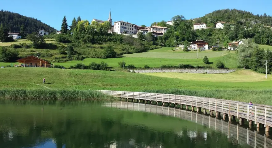 Una piacevole passeggiata dria al fos, con un ponte di legno che attraversa il lago e un villaggio sullo sfondo.