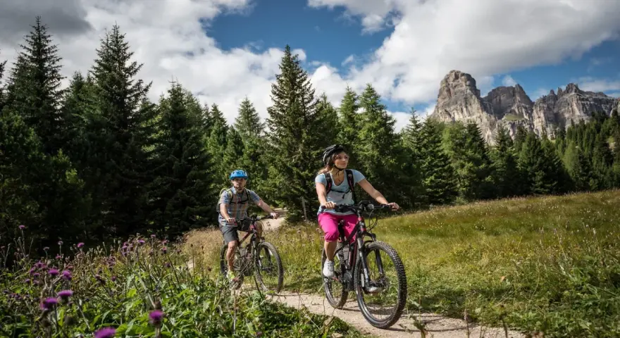 Coppia in mountain bike su sentiero a Passo Campolongo, con bosco di abeti e montagne sullo sfondo.