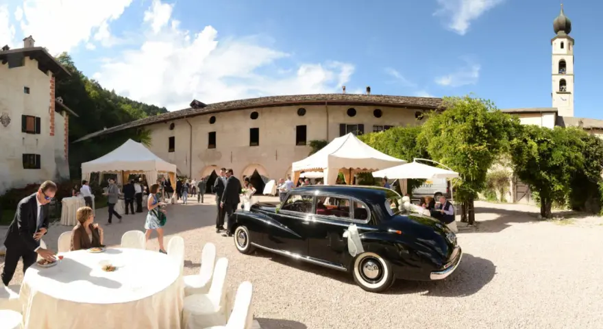 Matrimonio a Caldonazzo centro storico: auto d'epoca nera, tavoli allestiti, ospiti e un campanile sullo sfondo.