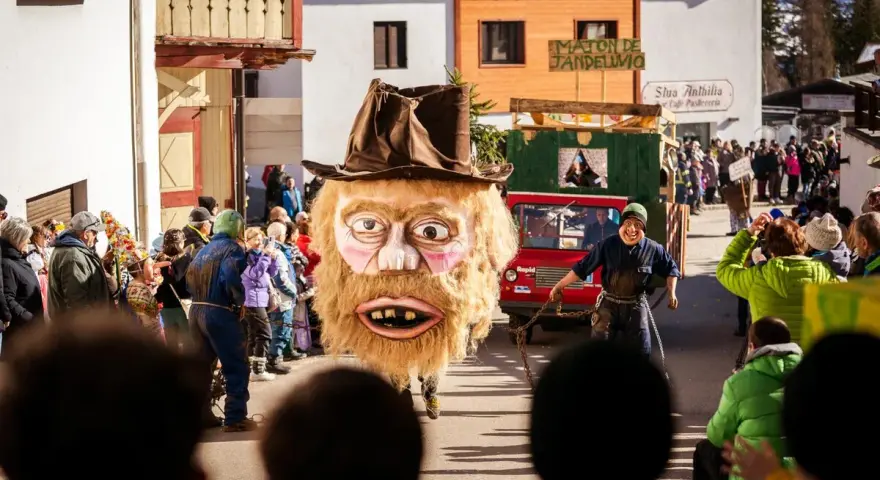 Gigantesca maschera pelosa con cappello marrone sfila al carnevale trentino. Un uomo con maschera grottesca trascina una catena.
