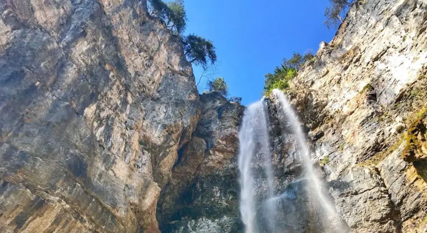 La cascata di Tret scende maestosa da una parete rocciosa imponente, sotto un cielo azzurro e limpido.