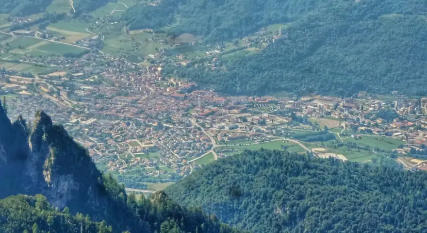 Vista aerea della Valsugana, Trento, con un paese incastonato tra montagne verdi e un picco roccioso in primo piano.