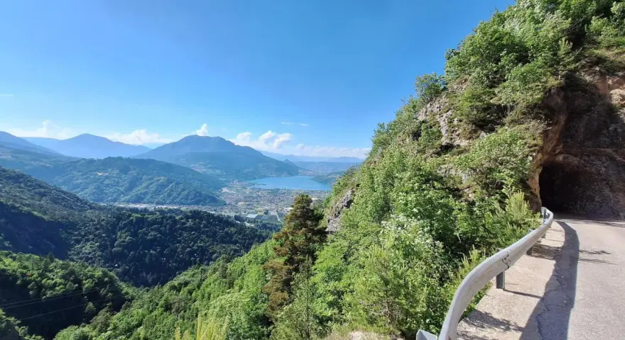 Vista panoramica dal Monte Vezzena con lago azzurro e montagne sullo sfondo. Strada con galleria scavata nella roccia.