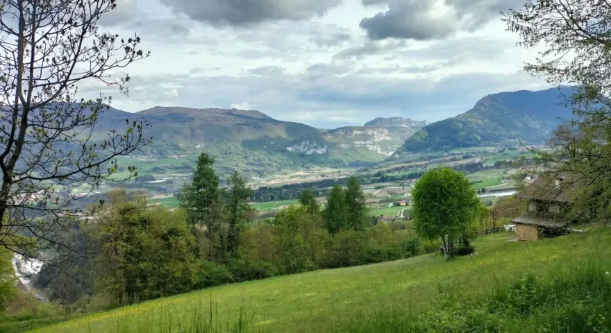 Prato verde e montagne sullo sfondo nella valle di Comano. Una casa con tetto in legno e un albero solitario.