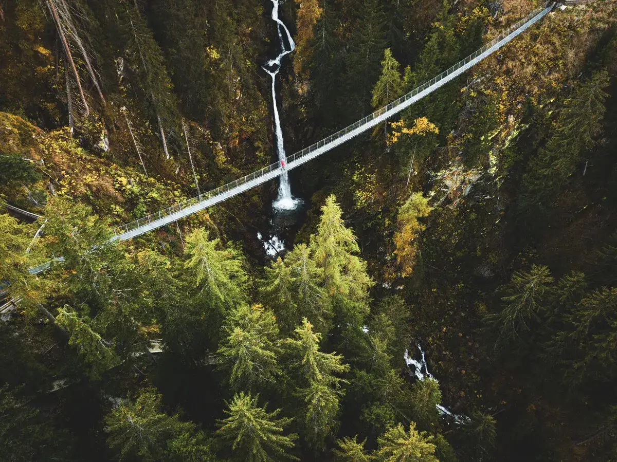Un uomo solitario attraversa un ponte tibetano sospeso su una cascata in una foresta autunnale.