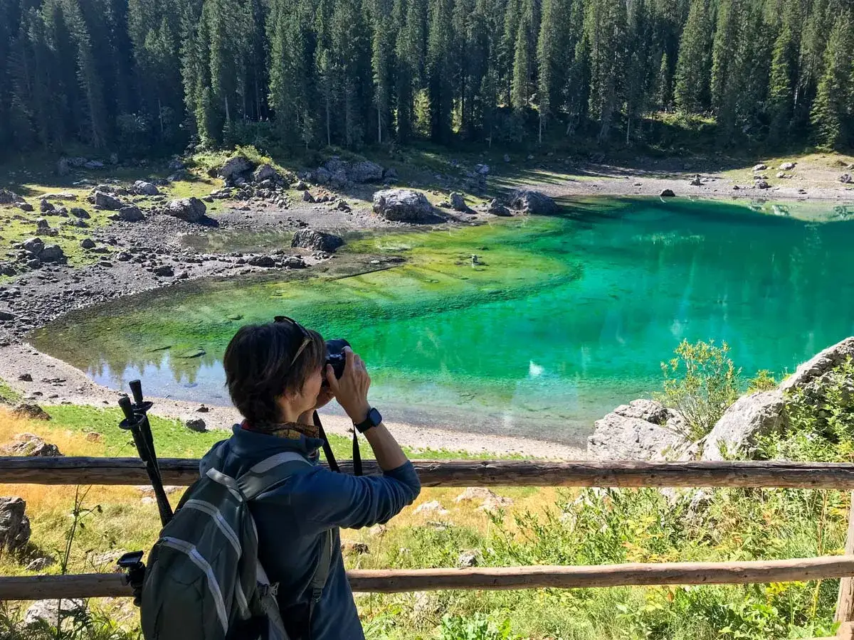 Donna con zaino e macchina fotografica immortalare un lago alpino dalle acque smeraldine. Un'esperienza di trekking facile e rigenerante.
