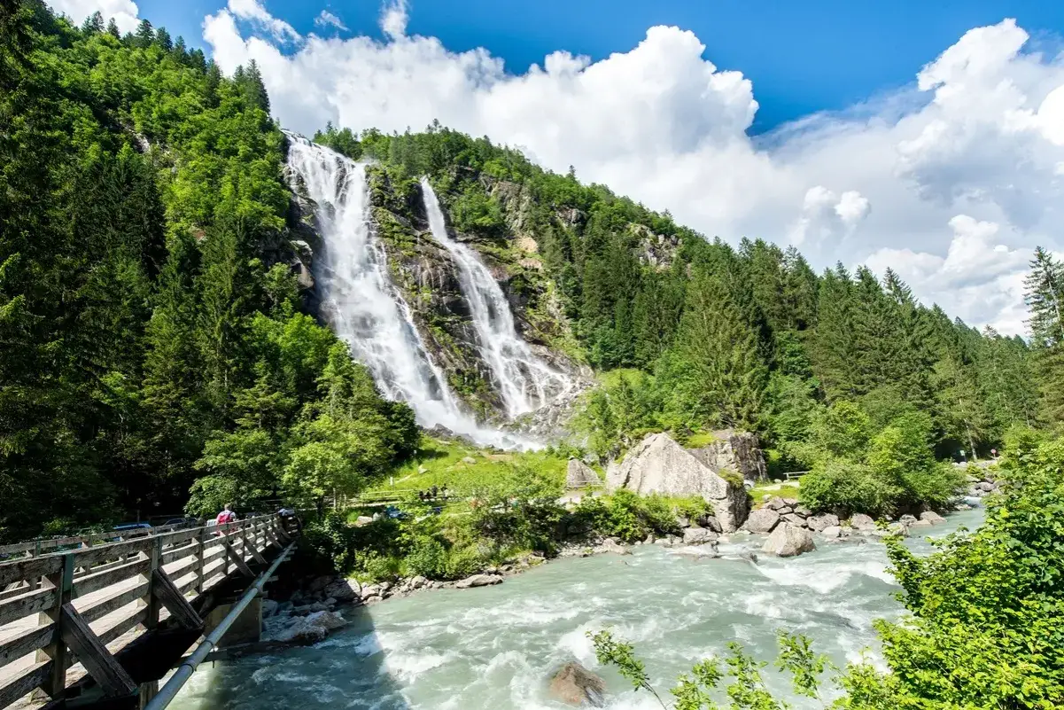 Cascata imponente nel Parco dell'Adamello, con un fiume impetuoso e un ponte di legno.
