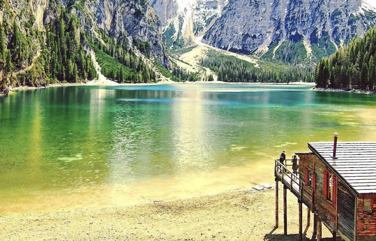 Lago alpino con acque smeraldine e montagne imponenti nel Trentino Alto Adige. Una baita di legno con due persone affacciate sul lago.