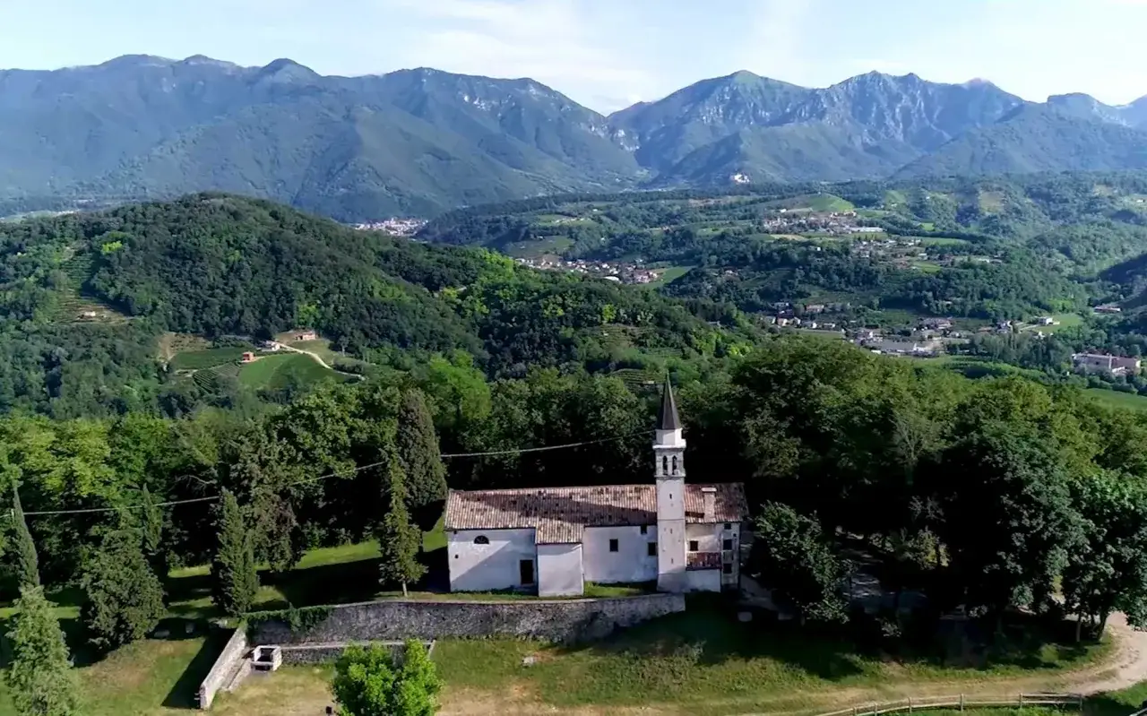 L'eremo di San Gallo, una chiesetta bianca con campanile, si erge tra alberi verdi con montagne sullo sfondo.