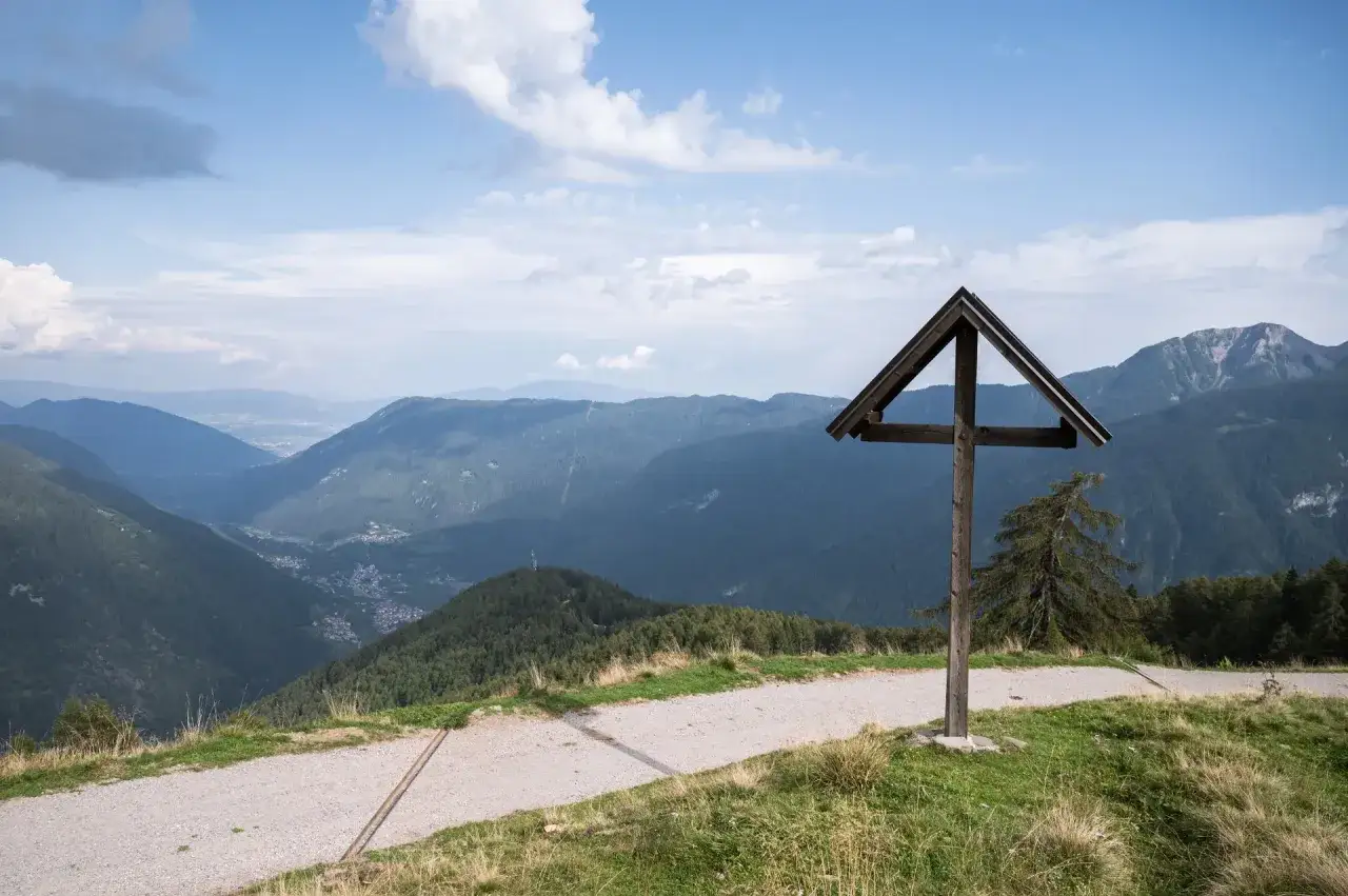 Croce in legno sul sentiero del Cimon di Bolentina, con vista su vallata alpina e cielo azzurro.