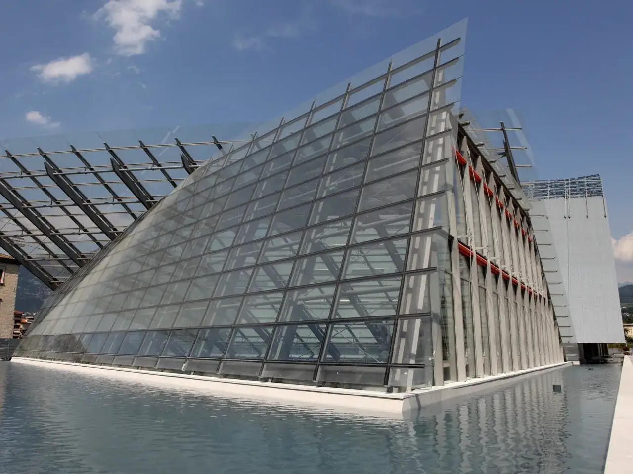 Il Museo delle Scienze di Trento si specchia in una vasca d'acqua, con la sua audace architettura in vetro e acciaio che cattura il cielo azzurro.