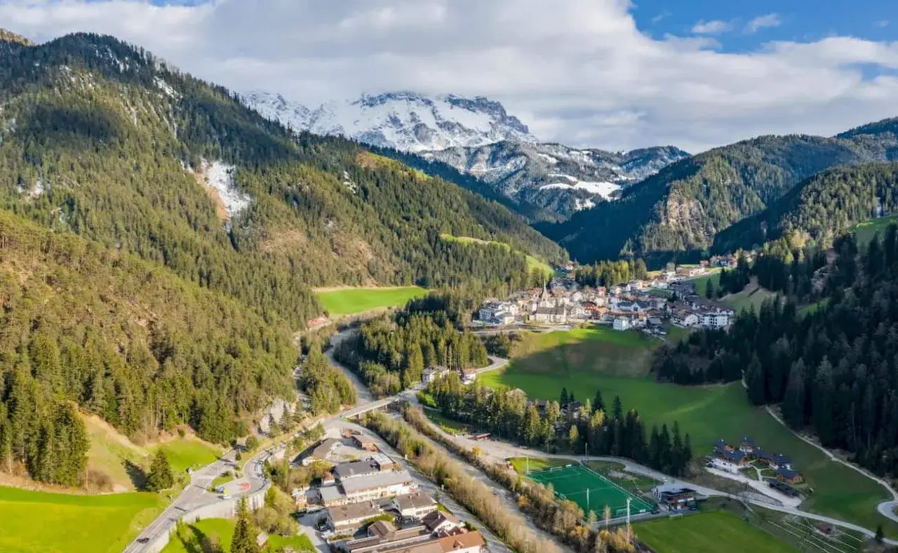 Villaggio incastonato tra verdi pendii e cime innevate nelle Alpi Giudicarie. Un fiume serpeggia tra boschi e prati, mentre un campo sportivo brilla al sole.