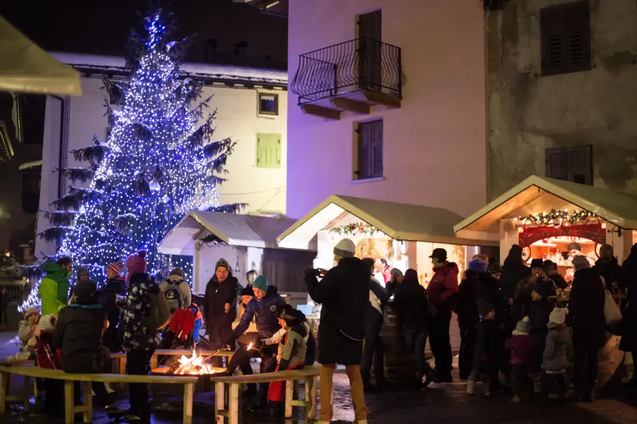 Atmosfera magica ai mercatini di Natale di Ossana, con un grande albero illuminato e gente riunita attorno a un fal&ograve;.