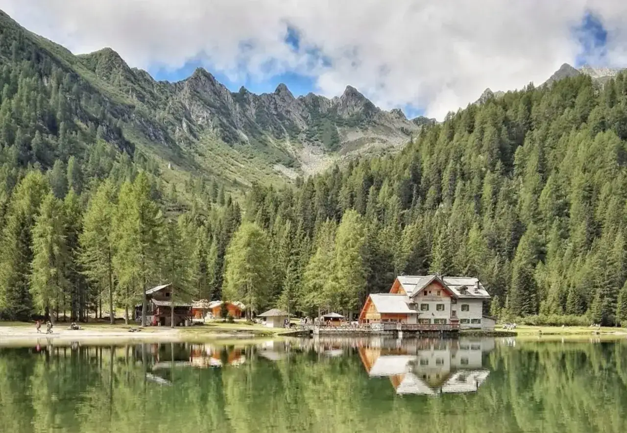 Riflessi di case e montagne nel lago di Madonna di Campiglio, circondato da fitti boschi di conifere.