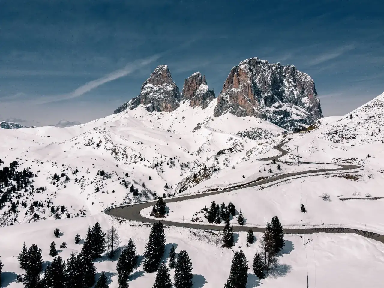 Strada tortuosa tra i passi delle Dolomiti innevate, con imponenti vette rocciose sullo sfondo e pini solitari.