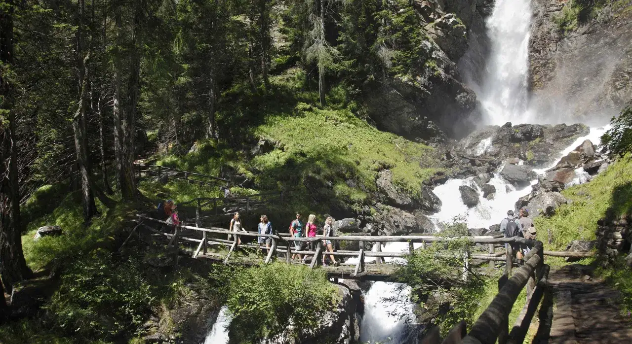 Un gruppo di escursionisti attraversa un ponte di legno sul sentiero del Val di Rabbi, con una cascata imponente sullo sfondo.