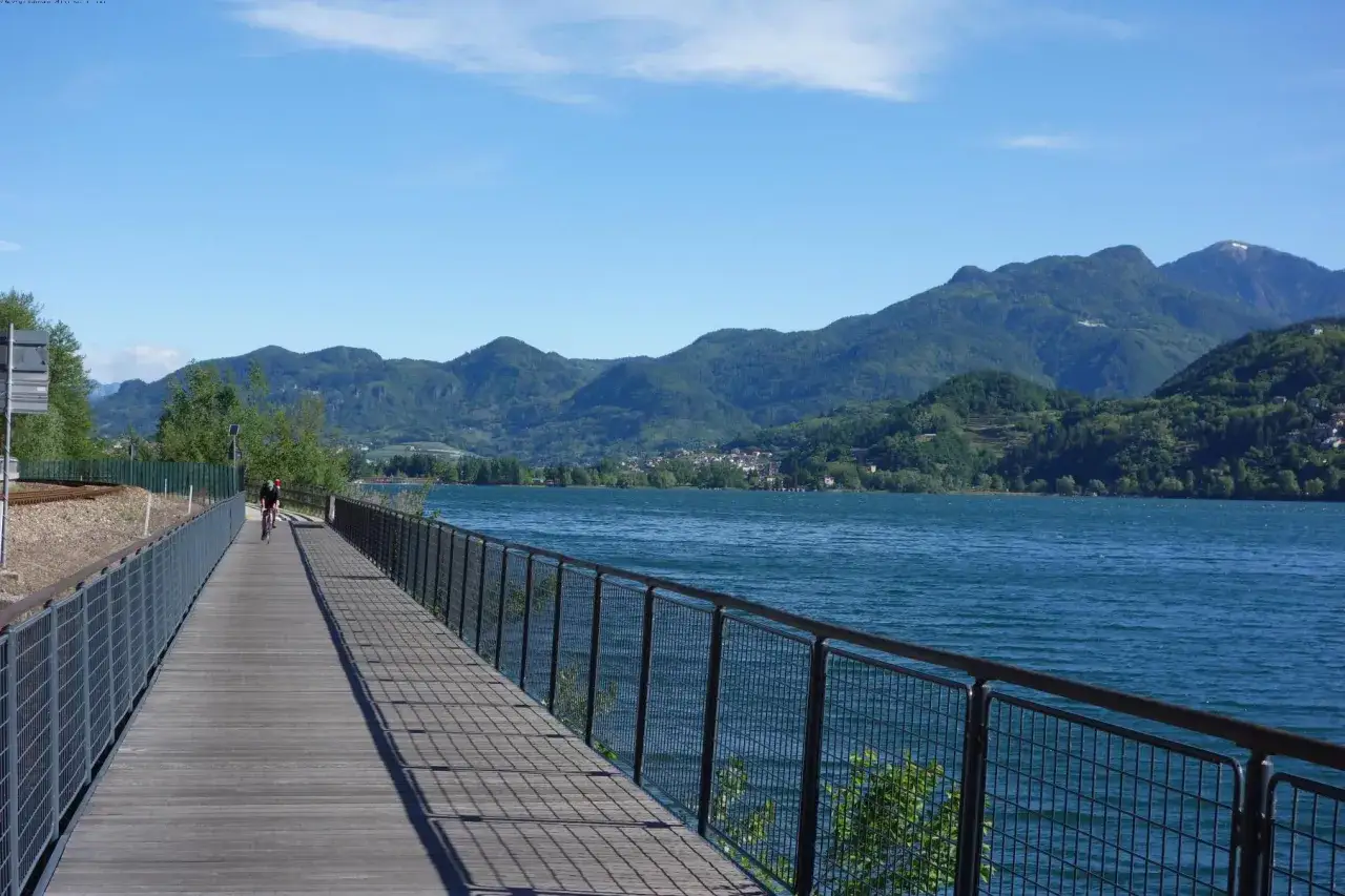 Ciclista su passerella in legno lungo lago blu, con montagne verdi sullo sfondo. Un'ottima idea per cosa vedere a Levico Terme.