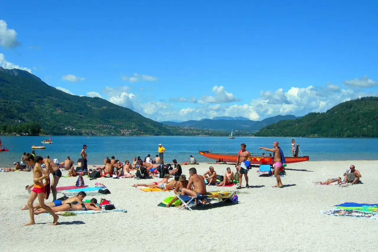 Gente in spiaggia e in acqua al lago di Caldonazzo, con montagne verdi sullo sfondo e un cielo azzurro con nuvole bianche.