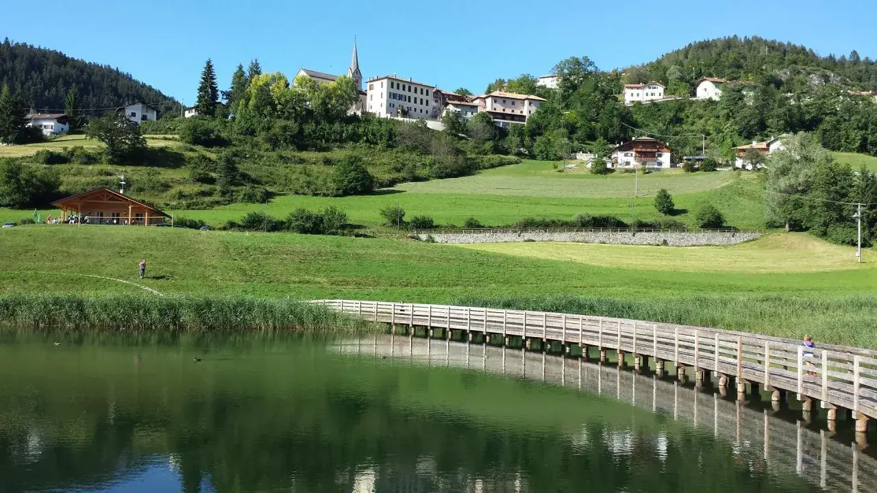 Passeggiata sul pontile dei laghetti di Ruffrè, con case e verdeggianti colline sullo sfondo.