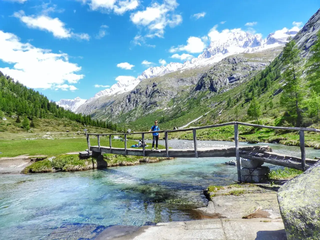 Un uomo su un ponte di legno nel Val di Fumo, con montagne innevate sullo sfondo e un fiume cristallino.