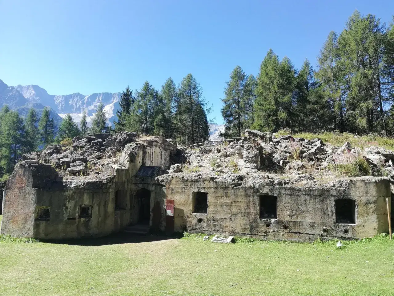 Rovine di un forte alpino, parte del giro dei forti tonale, con montagne innevate sullo sfondo e alberi verdi.
