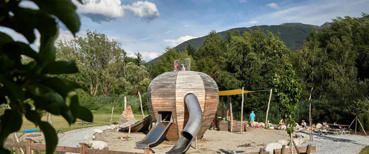 Un colorato parco giochi trentino con una struttura a forma di sfera in legno, scivoli e bambini che giocano.