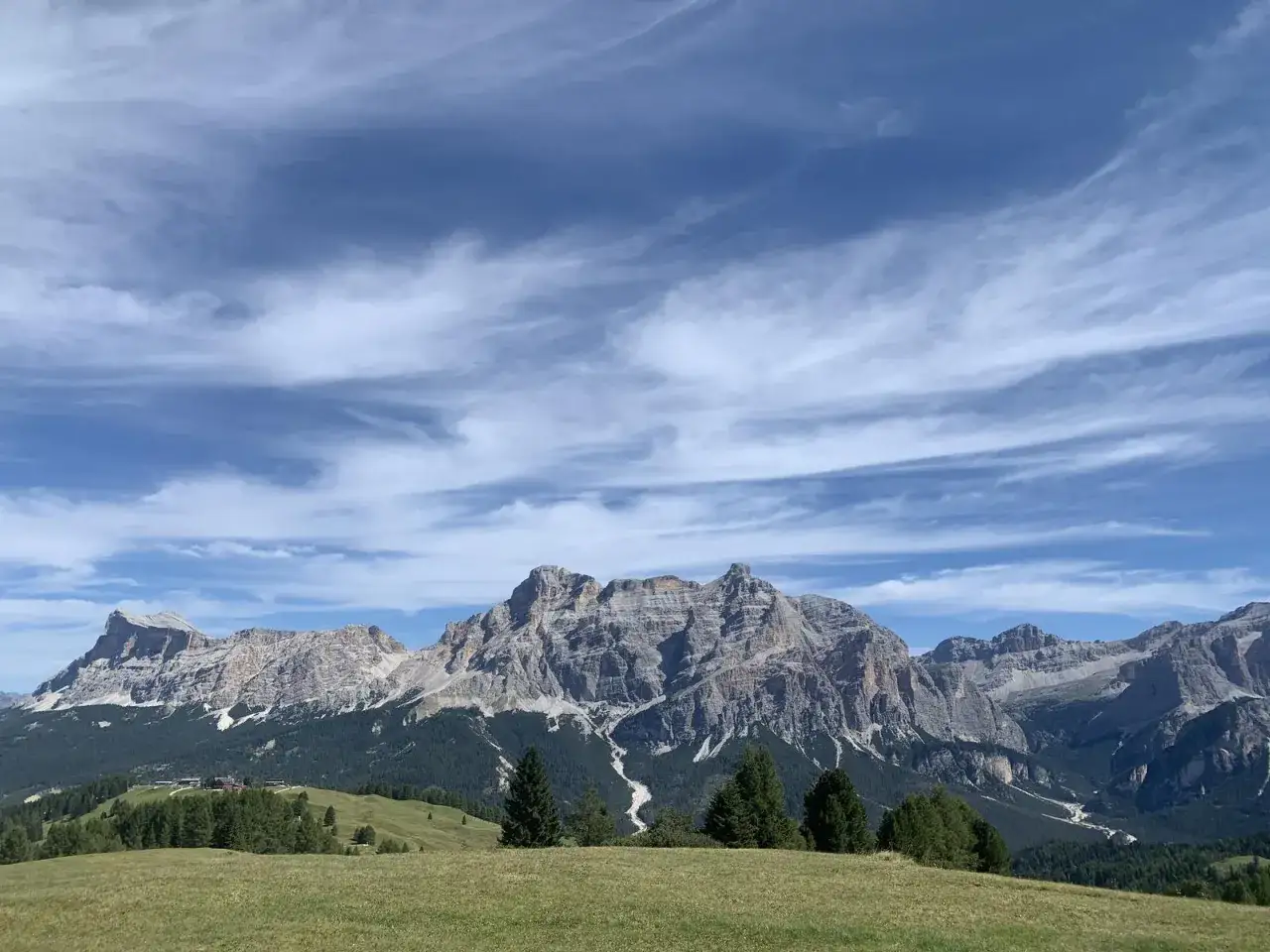 Panoramica mozzafiato delle Dolomiti dal Passo Campolongo, con cime rocciose, boschi verdi e un cielo azzurro striato di nuvole bianche.