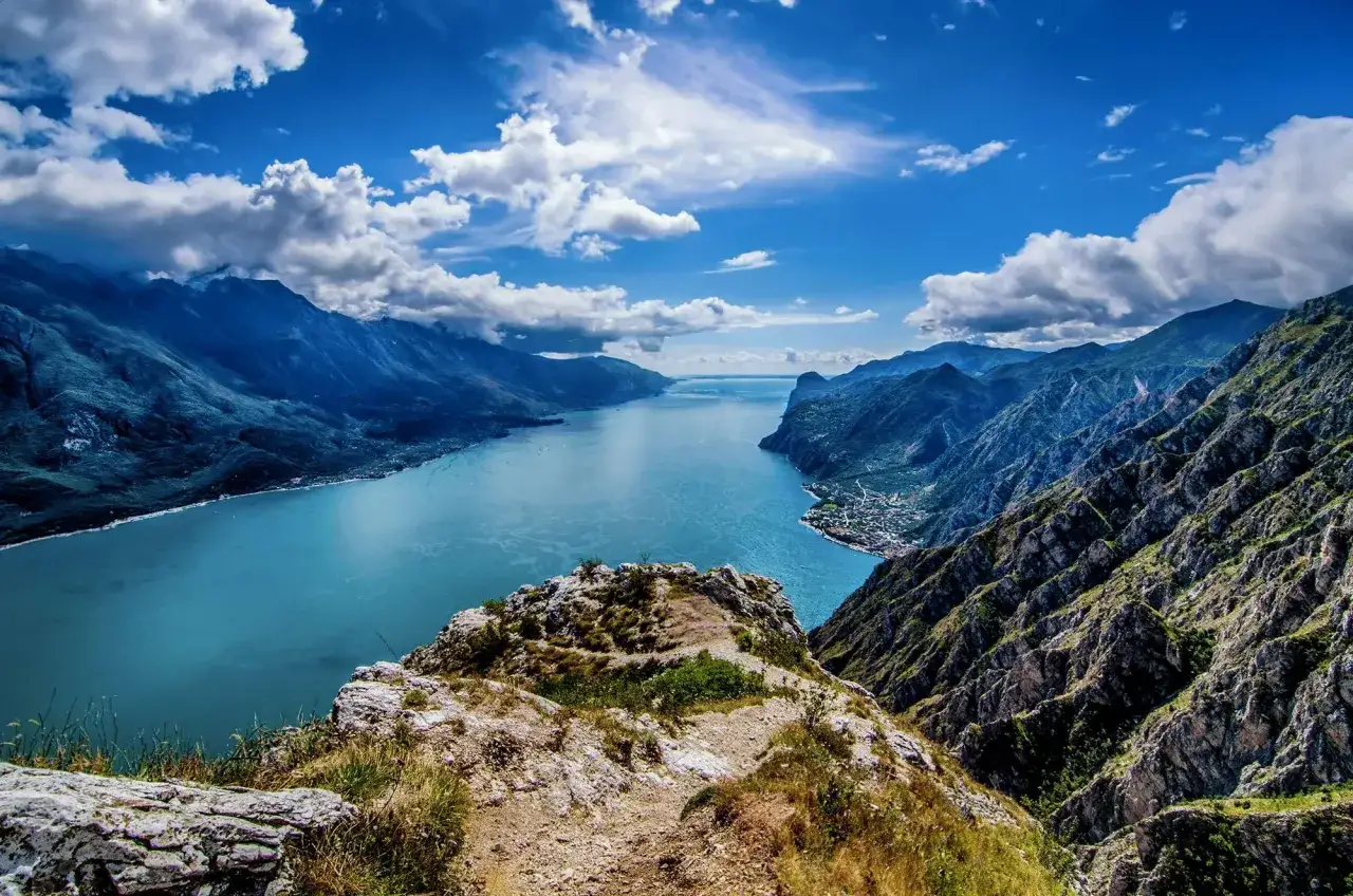 Vista mozzafiato da un sentiero per fare trekking, con un lago azzurro incastonato tra montagne scoscese e un cielo blu con nuvole bianche.