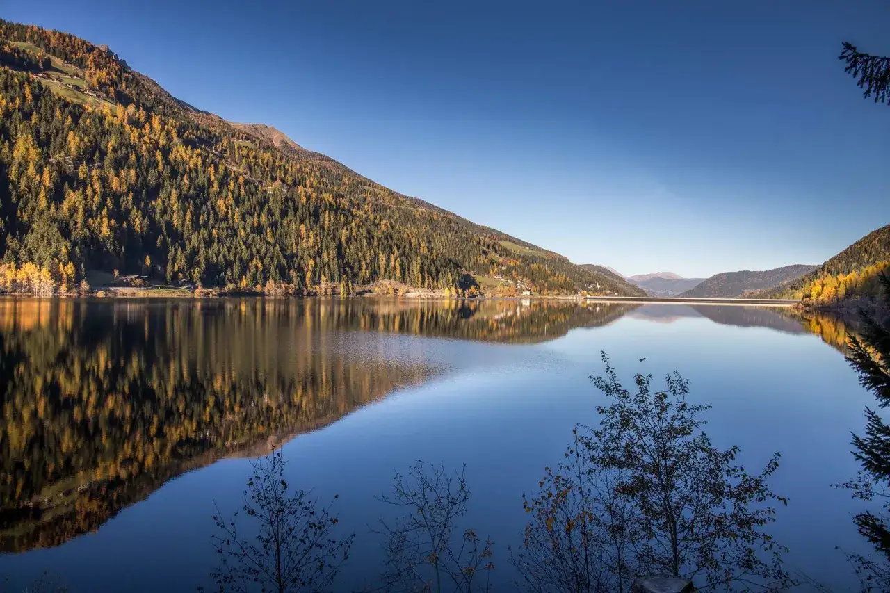 Autunno dorato sul Val d'Ultimo lago, con boschi che si specchiano nelle acque calme e un cielo terso.