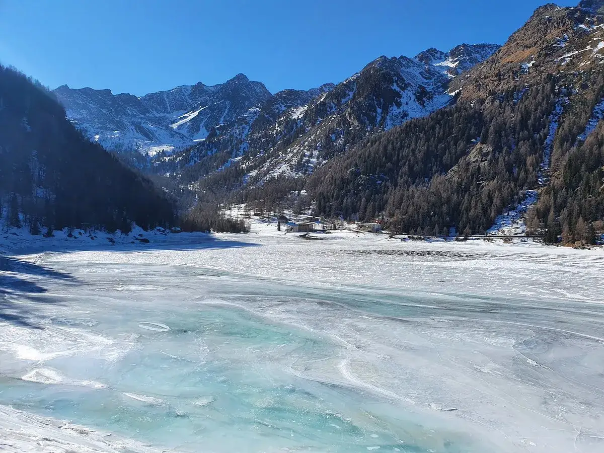 Il lago di Fontana Bianca, ghiacciato e scintillante sotto il sole, circondato da montagne innevate e boschi scuri.