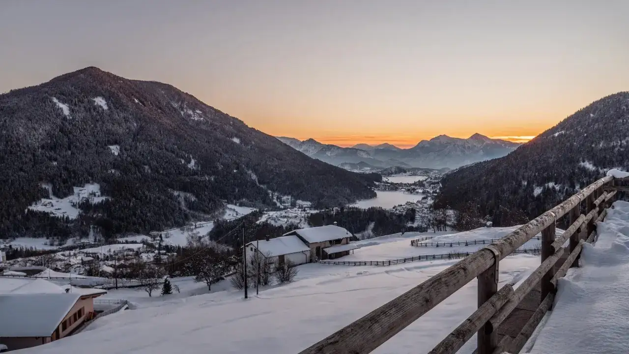 Alba innevata su Bedollo, Trento. Case e boschi coperti di neve, con montagne sullo sfondo e un cielo aranciato.