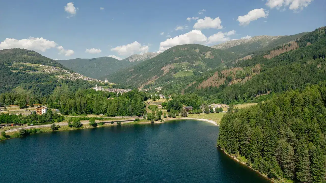 Il lago delle Piazze, incastonato tra montagne boscose, riflette il cielo azzurro. Un villaggio alpino si aggrappa al pendio verdeggiante.