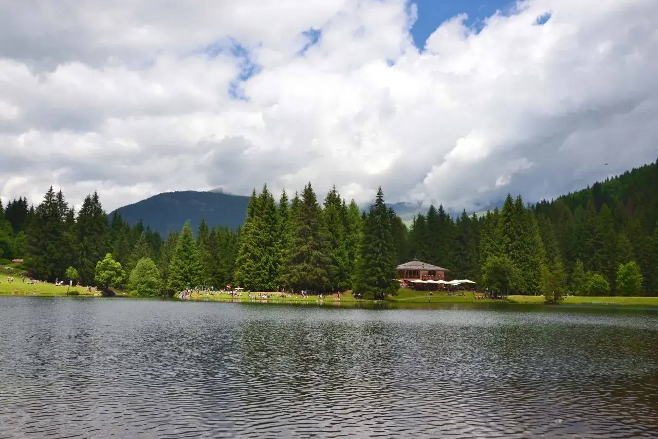 Laghi Val di Sole raggiungibili in auto: un lago tranquillo circondato da abeti, con persone che si rilassano sull'erba e un rifugio alpino.
