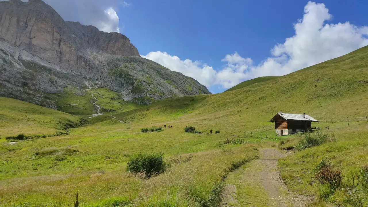 Un sentiero conduce a un rifugio alpino ai piedi del monte Catinaccio, tra verdi pascoli e un cielo azzurro con nuvole bianche.