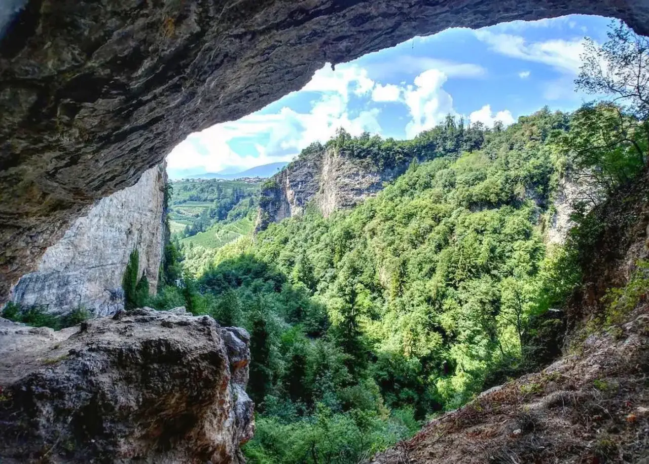 Vista da una grotta sul verdeggiante canyon Val di Non, con rocce imponenti e cielo azzurro.