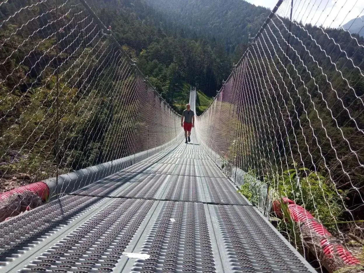 Un uomo cammina su un lungo ponte tibetano nelle Dolomiti, circondato da una foresta verdeggiante.