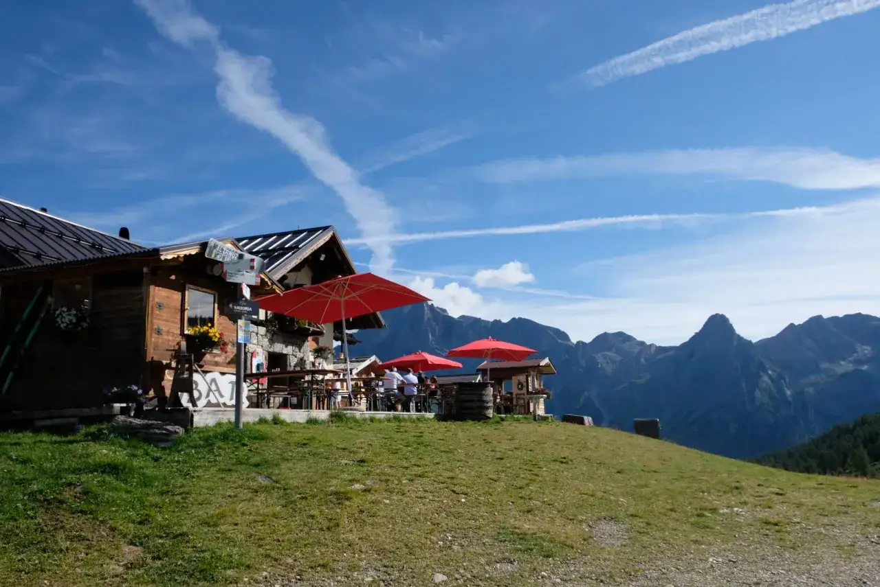 Rifugio alpino con tavoli all'aperto e ombrelloni rossi, sullo sfondo le vette maestose per il giro delle malghe San Martino di Castrozza.