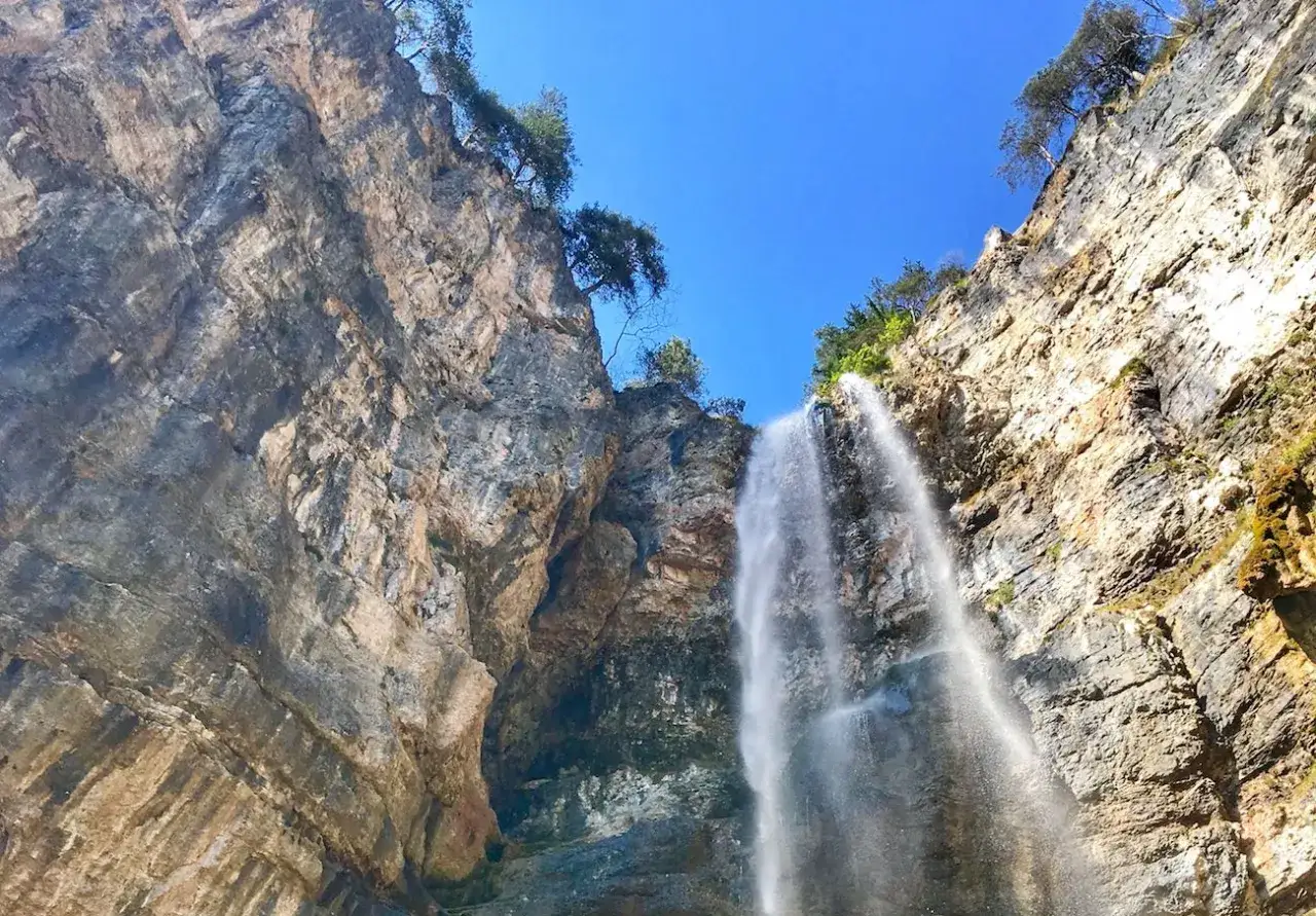 La cascata di Tret scende maestosa da una parete rocciosa imponente, bagnata da un cielo azzurro intenso.