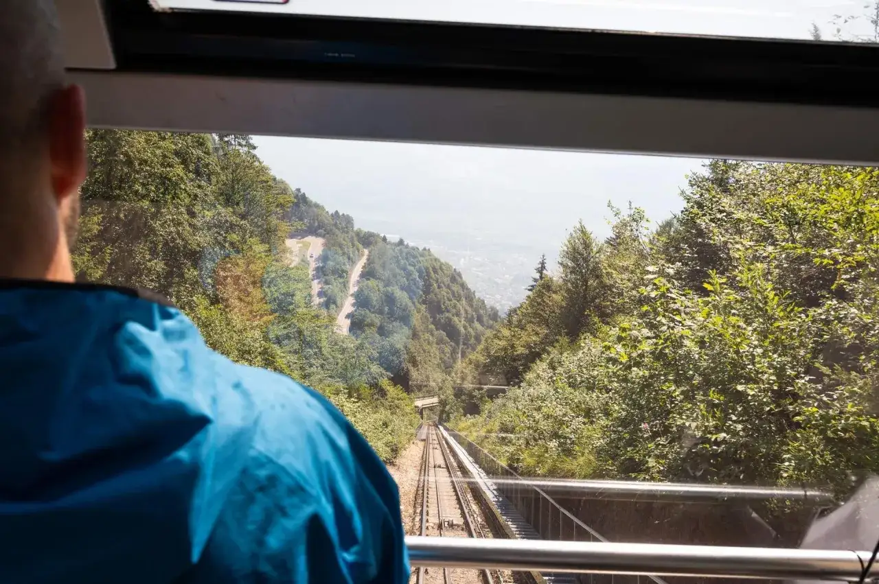 Vista dal vagone di una funicolare che scende verso valle, tra alberi rigogliosi. Si intravede una strada tortuosa e un paese in lontananza. Un viaggio verso il passo Mendola.