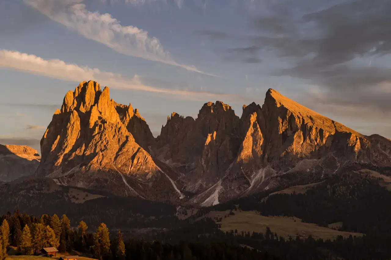 Un magico tramonto sulle Dolomiti illumina le cime rocciose con toni dorati, mentre nuvole leggere solcano il cielo.