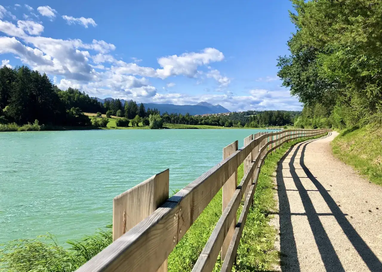 Sentiero lungo il lago dal colore turchese, con una staccionata in legno e montagne sullo sfondo. Un luogo incantevole, il tavon di Coredo.