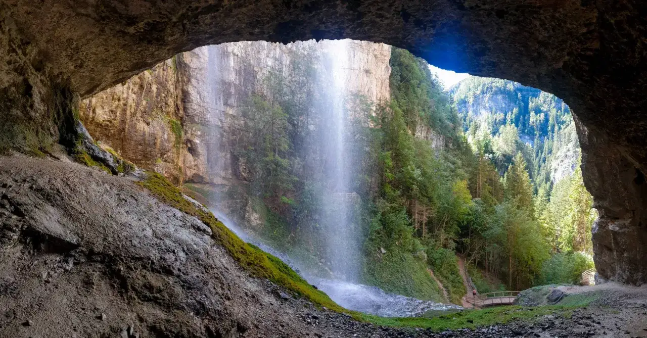 Vista da una grotta su spettacolari cascate del Trentino, con sentieri e ponticelli tra la vegetazione lussureggiante.
