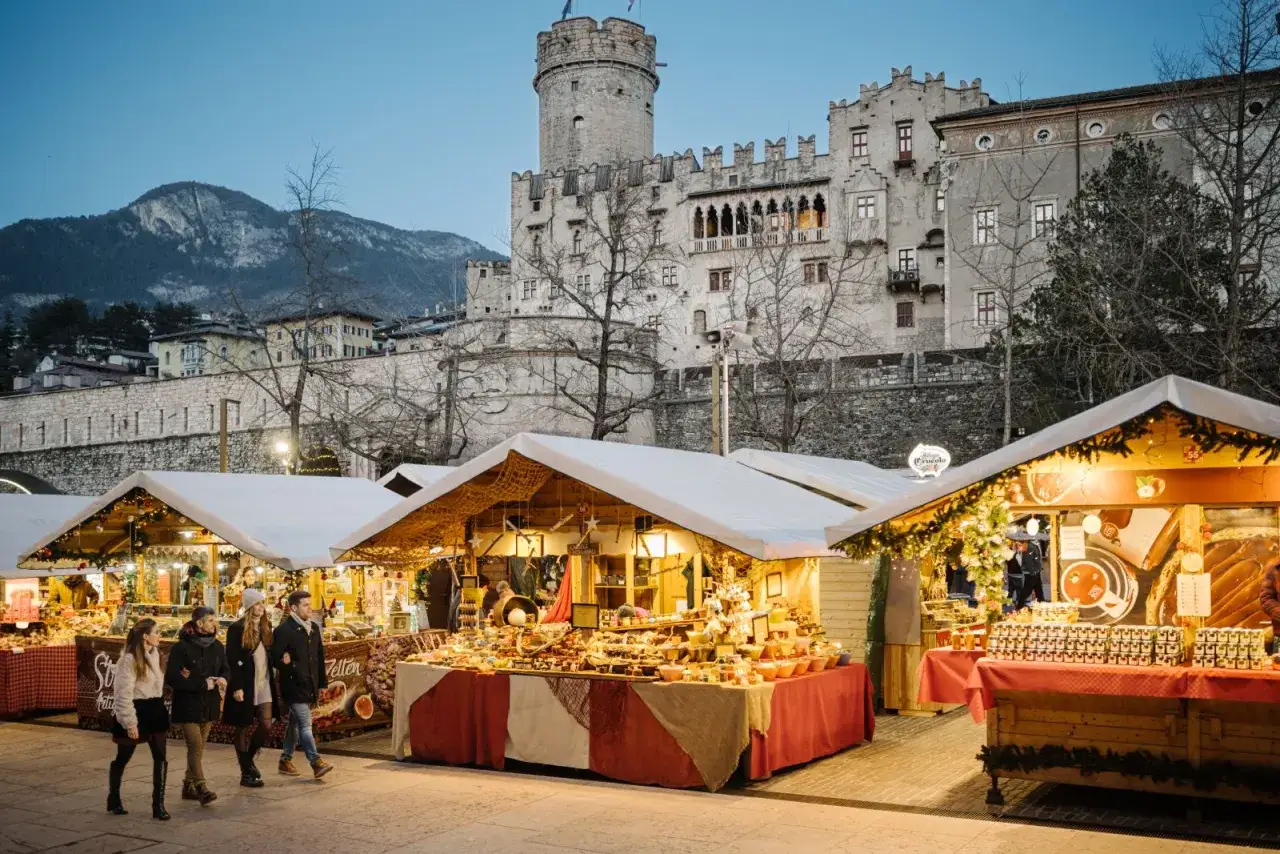 Mercatino di Natale a Trento con bancarelle illuminate e il castello sullo sfondo. Atmosfera magica.